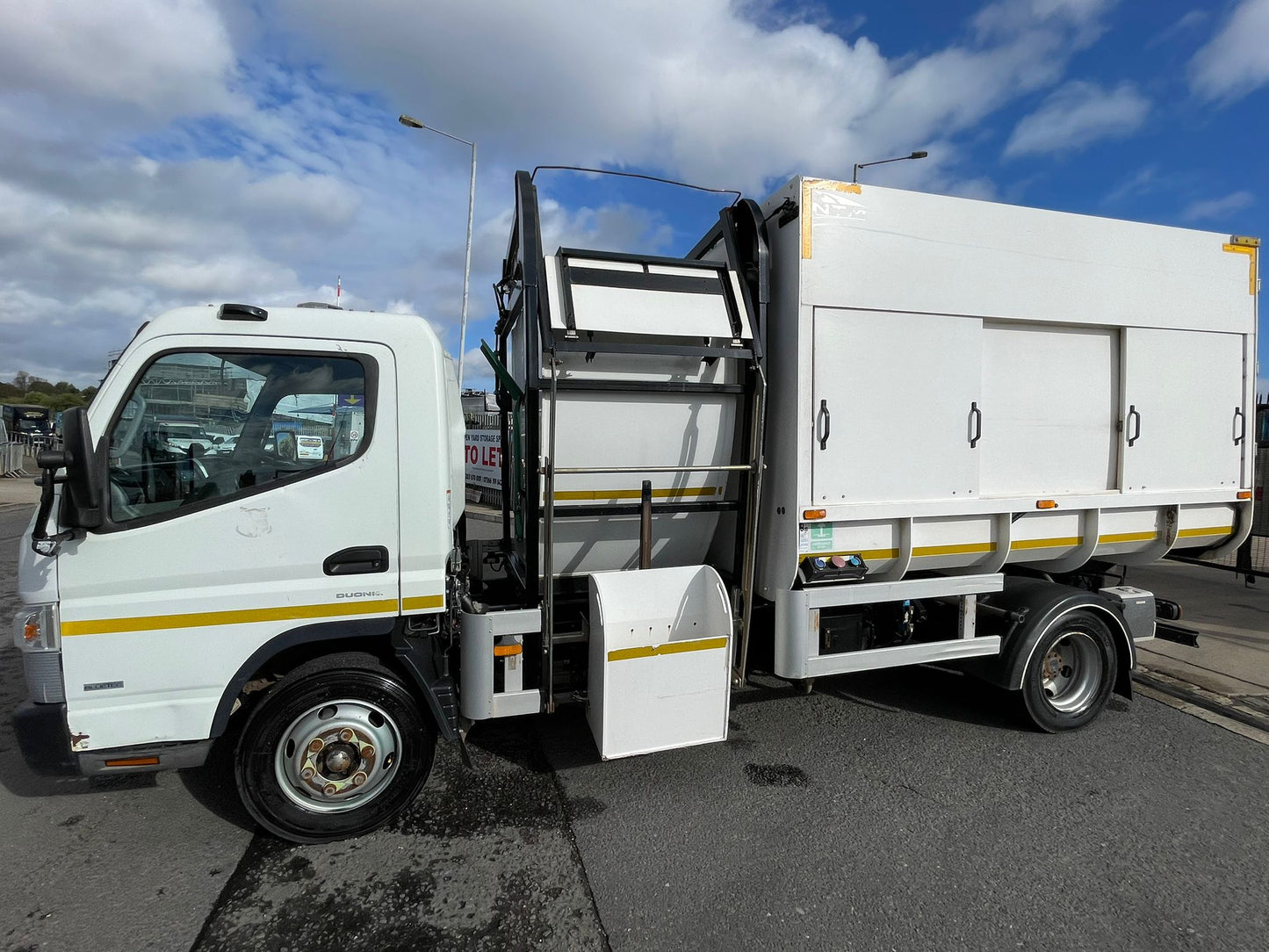 White garbage truck with open rear door on a road under a blue sky with clouds.